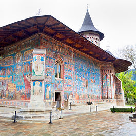 A ground-level view of the vibrant exterior frescoes of the Voroneț Monastery in Bukovina, Romania, famous for its intense shade of blue ("Voroneț Blue"). The medieval Orthodox church features detailed religious murals covering its outer walls, protected by a wide overhang with a steep wooden-shingled roof and a pointed spire, set on paved stone grounds surrounded by green trees.