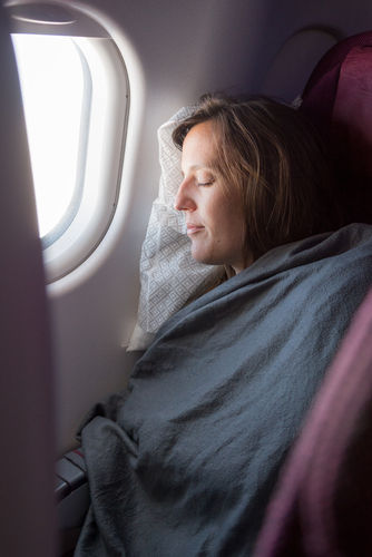 A side profile of a woman with brown hair, eyes closed, is shown sleeping comfortably in an airplane seat. She is covered with a dark grey blanket and her head rests on a patterned light grey pillow against the seat's headrest. Bright sunlight streams in through the airplane window to her left, illuminating her face. The seat's reddish-purple upholstery is visible on the right.