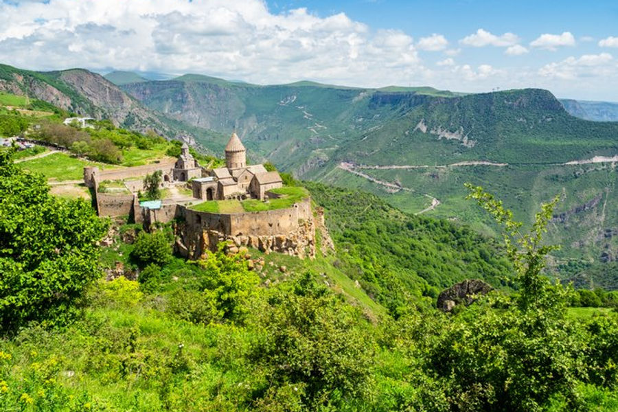 A wide landscape view of the medieval stone Tatev Monastery perched high on a cliff edge above a deep, lush green mountain gorge under a partly cloudy sky.