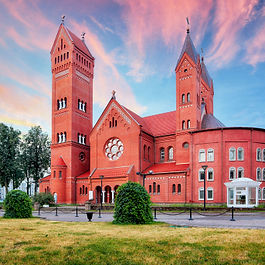 A vibrant sunset view of the Church of Saints Simon and Helena, popularly known as the Red Church, located near Independence Square in Minsk, Belarus. Built in Neo-Romanesque style, the striking red-brick Roman Catholic church features a large central rose window, prominent rectangular and pointed spires, and manicured green lawns under a soft, pink-and-blue streaked sky.