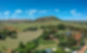 A scenic view of a green, hilly landscape under a clear blue sky with scattered clouds. In the foreground, there are cultivated fields, trees, and several buildings with tin roofs, surrounded by lush vegetation. A prominent, tree-covered hill rises in the midground, set against a backdrop of more distant hills.