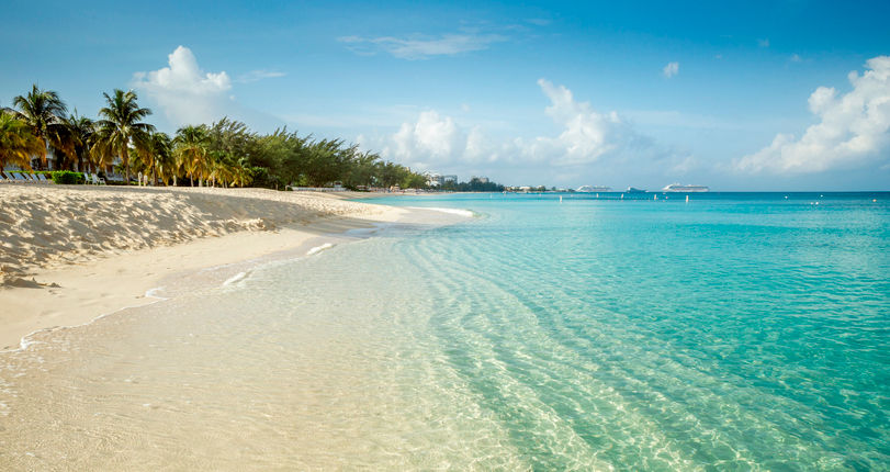 A wide-angle view of the pristine Seven Mile Beach in Grand Cayman, featuring crystal-clear turquoise water gently lapping onto soft white sand. Tropical palm trees line the shore under a bright blue sky with scattered white clouds, and cruise ships are visible on the distant horizon.