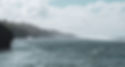 Coastal view of Atiu in the Cook Islands, featuring misty sea cliffs, gentle waves, and a forested shoreline under a partly cloudy sky, evoking a sense of serene remoteness.