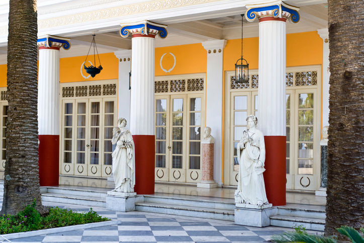 Classical colonnaded porch of the Achilleion Palace featuring neoclassical statues, painted columns, and checkerboard floor tiles in Corfu.