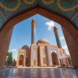 A symmetrical view framing the Imamzadeh Complex in Ganja, Azerbaijan, through a large brick pointed arch adorned with colorful blue floral tile mosaic patterns. The complex features prominent red-brick architecture, turquoise-tiled domes, and tall, spiraled minarets with blue tiled accents standing under a clear blue sky.