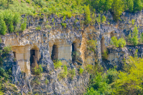 A high-angle view of a steep cliffside made of light-colored limestone with large cave openings carved into the rock, surrounded by lush green foliage.
