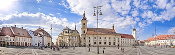 A wide panoramic shot captures the Grand Square in Sibiu, Romania, bustling with people under a bright blue sky with fluffy white clouds. The square is paved with cobblestones, and is surrounded by numerous historic buildings of various architectural styles and colors. Prominent features include a tall clock tower and a large, light-colored church building, along with decorative lampposts. The sun is shining brightly from the upper left, casting light on the scene.