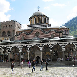 A full view of the main church at Rila Monastery in the Rila Mountains, Bulgaria. Tourists walk across the wide cobblestone courtyard in front of the eastern Orthodox church, which features distinctive black-and-white striped arches, colorful frescoed porticos, a central dome topped with a cross, and the ancient stone Hrelyo Tower standing beside it under a partly cloudy sky.