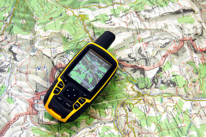 A bright yellow and black handheld GPS device is positioned diagonally on top of a detailed topographical map. The GPS screen shows a digital map display. The background map, primarily in shades of green, white, and brown with contour lines and labels, suggests a mountainous or hilly terrain. The GPS device is clearly in focus, while the map beneath it is slightly blurred, emphasizing the device as the subject.