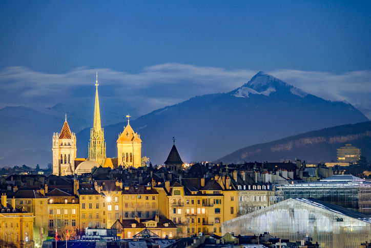 A stunning twilight skyline view of Geneva, Switzerland, with the historic city center illuminated by warm streetlights. The prominent stone towers and tall, slender green spire of St. Pierre Cathedral rise above a dense cluster of classic European buildings. In the background, a massive, dark mountain with a snow-dusted, jagged peak stands sharply against a deep blue evening sky filled with low, soft clouds.