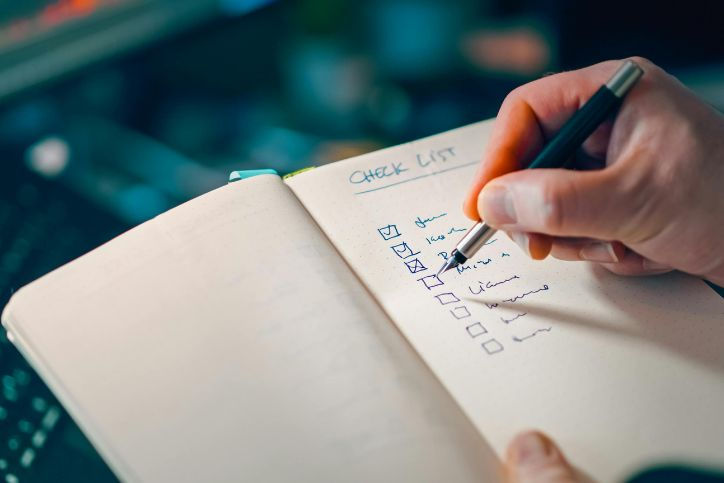 A close-up, shallow-focus shot of a person’s hand using a silver pen to check off items in a notebook. The page is titled "CHECK LIST" in blue ink, with several boxes already marked with an "X."