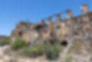 Ruins of the Ingurtosu Mine in Sardinia, Italy, featuring crumbling stone and brick walls, exposed structural beams, and overgrown vegetation under a clear blue sky, reflecting the area's industrial heritage.