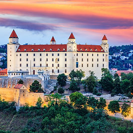 A grand sunset landscape view of Bratislava Castle (Bratislavský hrad) in Slovakia. The massive rectangular white palace with four corner towers and red-tiled roofs stands atop a hill above the Danube River, framed by illuminated stone fortifying walls and lush green trees beneath a brilliant orange, pink, and purple evening sky.