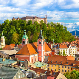 An elevated cityscape view of Ljubljana, Slovenia. In the foreground, terracotta-tiled roofs, colorful pastel buildings, and green-copper church steeples line the old town. In the background, medieval Ljubljana Castle sits atop a lush, forest-covered hill under a bright blue sky filled with soft white clouds.