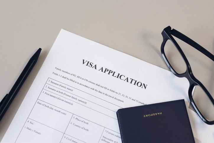 Close-up of a visa application form on a desk, accompanied by a black pen, a pair of eyeglasses, and a dark-colored passport—representing the process of preparing travel documents for international travel.