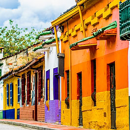 A colorful street-level perspective of Spanish colonial architecture in the historic La Candelaria district of Bogotá, Colombia. Boldly painted facades in vibrant hues of orange, yellow, purple, and green line a cobblestone pavement, adorned with traditional wooden balconies, tiled roofs, and window grilles against a backdrop of distant green hills and cloudy blue skies.