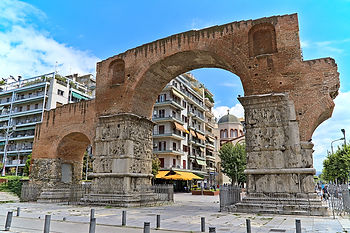 The Arch of Galerius in Thessaloniki, Greece, an ancient Roman structure made of brick and stone with detailed carved reliefs on its marble columns. The arch stands prominently in a modern urban setting, surrounded by apartment buildings, shops, and a nearby church dome visible in the background.