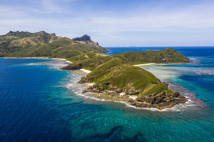 High-angle aerial view of a narrow, hilly green peninsula with coral reefs fringing clear turquoise and deep blue waters in the Yasawa Islands.