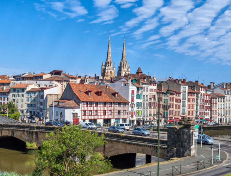 A wide shot of Bayonne, France, featuring the stone Pont du Génie bridge crossing the Nive river. In the background, the twin spires of the Bayonne Cathedral rise above a row of traditional Basque buildings with colorful timber-framed facades under a blue sky with wispy clouds.