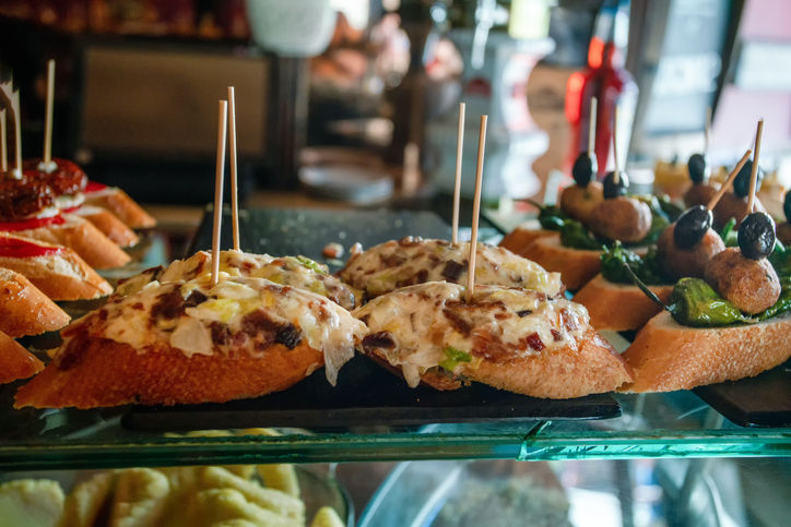 A close-up display of Spanish tapas, featuring toasted bread slices topped with melted cheese, bacon, and other savory ingredients, held with toothpicks, alongside other pintxos like olives and meatballs.