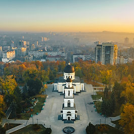 An elevated aerial shot looking down over the Metropolitan Cathedral of Christ's Nativity and its standalone bell tower in central Chișinău, Moldova. The neoclassical white church and plaza are surrounded by vibrant golden autumn trees, with the soft warm light of sunrise illuminating the urban skyline stretching into the hazy distance.