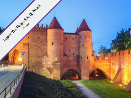 The image features the Warsaw Barbican, a historic fortified gate in Warsaw, Poland. It is depicted at dusk or night, illuminated by warm lights that highlight its red-brick construction and the two prominent conical-roofed towers. The Barbican has arched gateways and connects to a city wall on the right. In the foreground, there's a paved area and some greenery. A purple banner in the top left corner reads "Explore The Unique Side Of Poland."