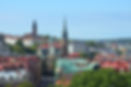 An elevated panoramic view showcases the city of Gothenburg, Sweden, under a clear blue sky. In the midground, a prominent red-brick church with a tall, slender green spire and a clock face dominates the skyline. To its left, another church with a reddish-brown brick tower is visible in the distance. The city's buildings in the foreground and midground feature a mix of red, orange, and green rooftops, with green trees interspersed throughout the urban landscape. In the far distance, the city stretches towards the horizon, with more buildings and greenery.