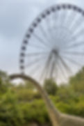 A unique outdoor scene features a large Ferris wheel rising behind a towering dinosaur statue. The Ferris wheel, a prominent feature against a cloudy sky, has numerous passenger gondolas. In the foreground, the long neck and head of a light-brown, textured dinosaur statue, resembling a Brachiosaurus, extends upwards from behind a dense line of green trees. The dinosaur's head is looking towards the left. More trees are visible between the dinosaur and the base of the Ferris wheel.