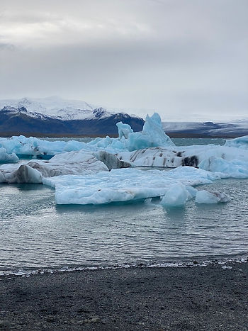 Floating icebergs drift through the glacial waters of Jökulsárlón Glacier Lagoon in Iceland, with snow-capped mountains rising in the background under a cloudy, overcast sky.