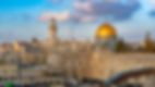 A sunset view of the Western Wall plaza in the Old City of Jerusalem, showing the light-colored stones of the Western Wall and the prominent golden Dome of the Rock and an adjacent minaret rising above the city skyline against a blue sky with pink-tinged clouds.