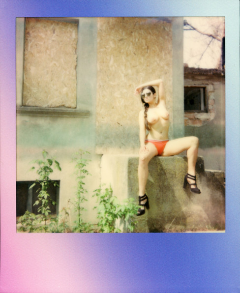A Polaroid of Misuzu topless in sunglasses and heels and sunglasses sat on steps of a boarded up building