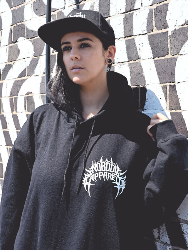 Misuzu wearing a black Nobody Apparel hoodie and snapback next to a black and white building