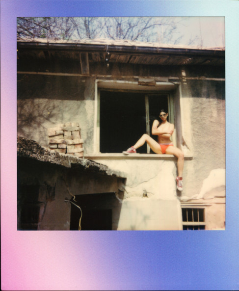 A Polaroid of Misuzu topless wearing sunglasses and converse, sat in a window of a derelict building