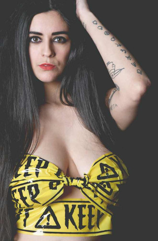 Portrait of Misuzu wearing yellow tape as a top with the words 'KEEP OUT' repeated. Her left hand is in her hair and she is staring straight at the camera