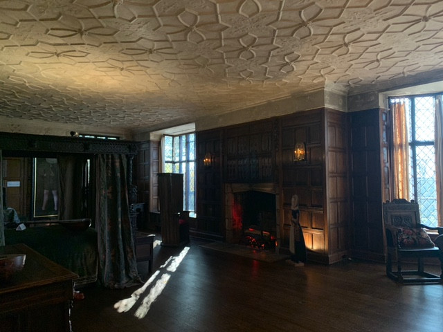 A large room with plaster ceilings with decorative geometric designs and dark wood panelling. Two windows frame a fireplace. There is a large bed draped in heavy fabric to the left in the image.