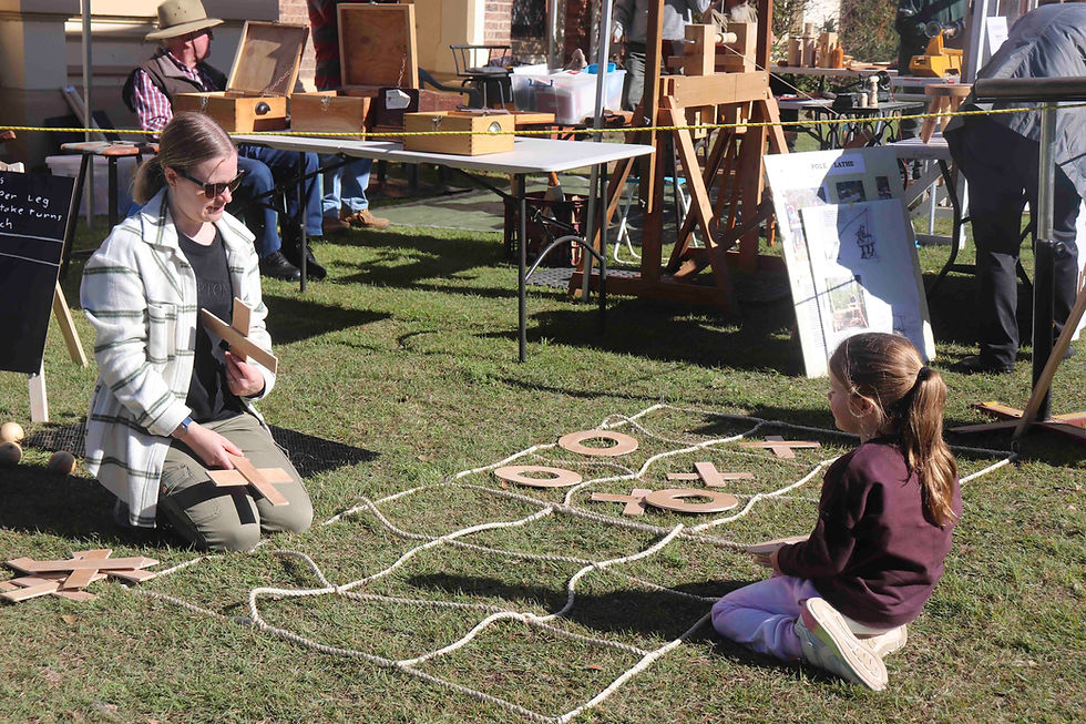 Woman and girl play tic-tac-toe on grass with wooden pieces. Background features tables with crafts. Sunny and relaxed setting.