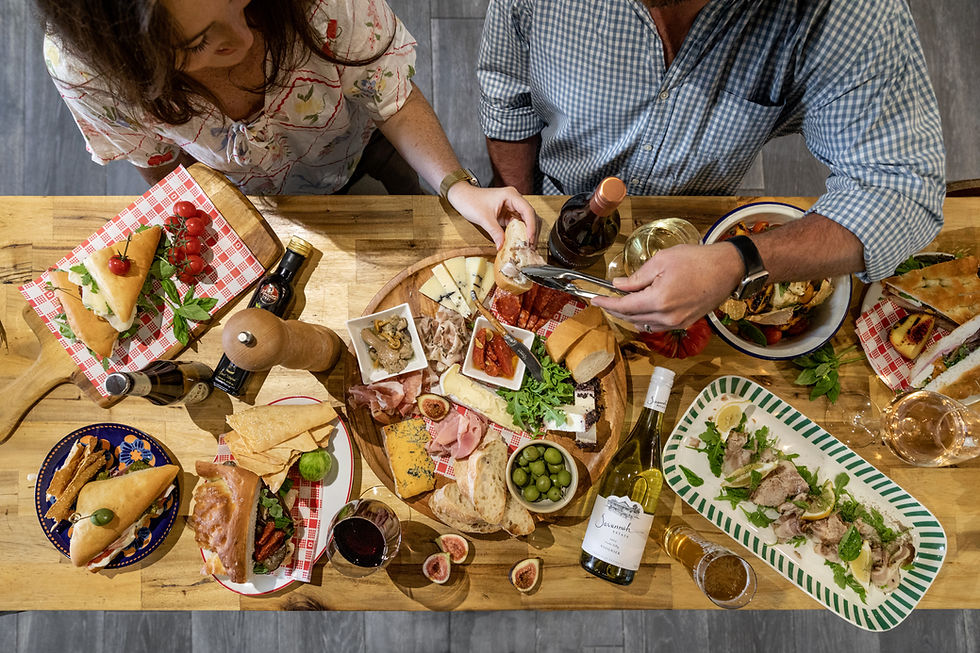 Overhead view of a couple serving themselves from a lavish table of cheeses, breads, olives, sandwiches and wine.