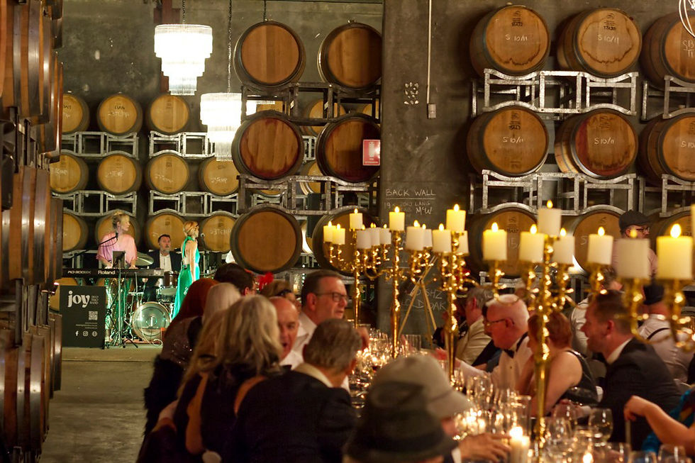 Guests dine at a long table with candlelight in a wine cellar. A band plays near stacked barrels. Warm, elegant ambiance.