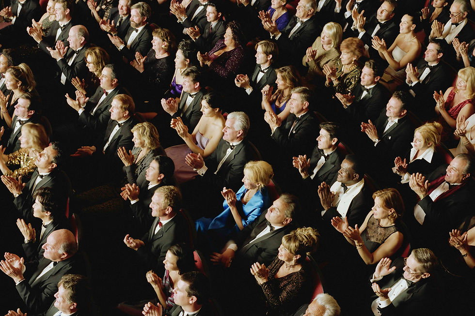 Audience in formal evening wear applauds in a dim theater, seated in red chairs.