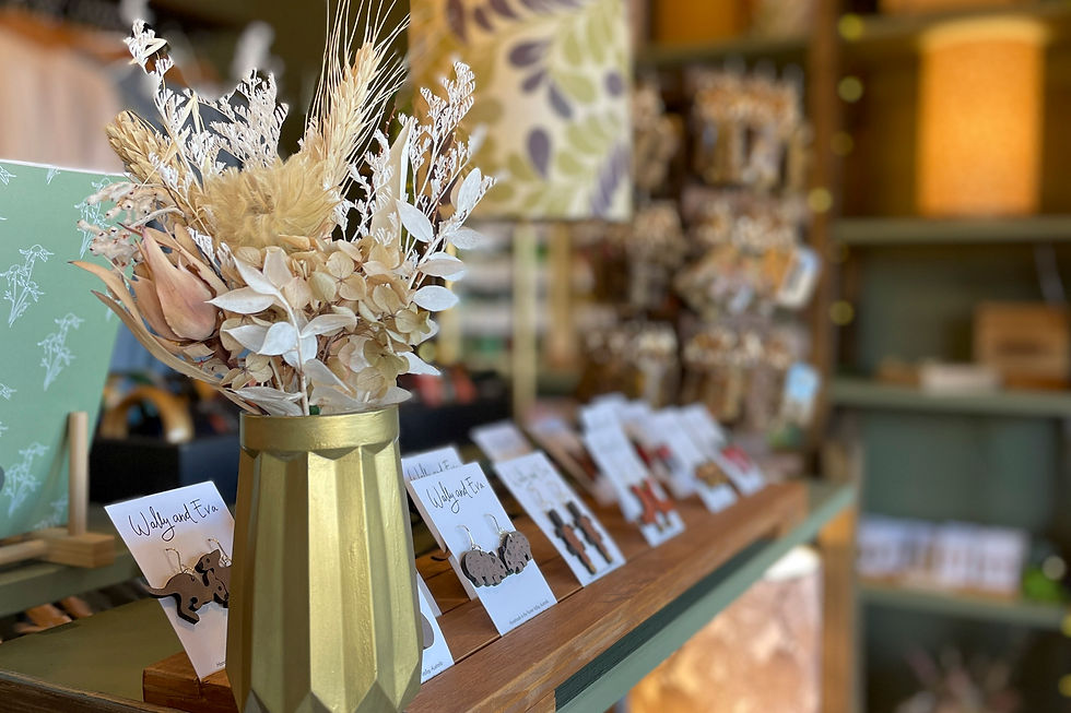 Golden vase with dried flowers on a wooden counter, surrounded by jewelry cards labeled "Wally and Eva." Blurred patterned backdrop.