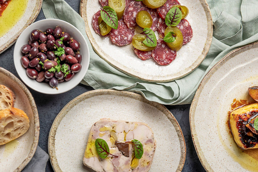 Plates of charcuterie with sliced meats, olives, and bread on a gray table. Garnished with herbs on a light cloth. Rustic and appetizing.