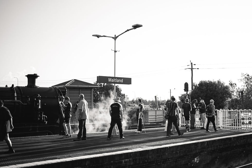 People gather on a train platform labeled "Maitland," near a steam locomotive. The scene is in black and white, with a relaxed mood.