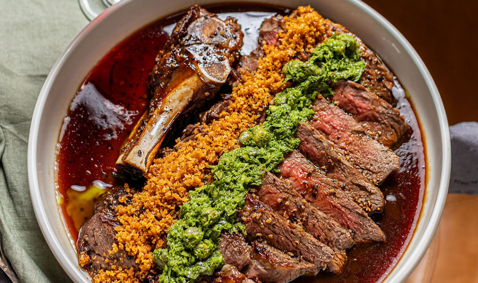 Sliced steak and bone marrow in a bowl, topped with green herb sauce and golden crumbs over red sauce.