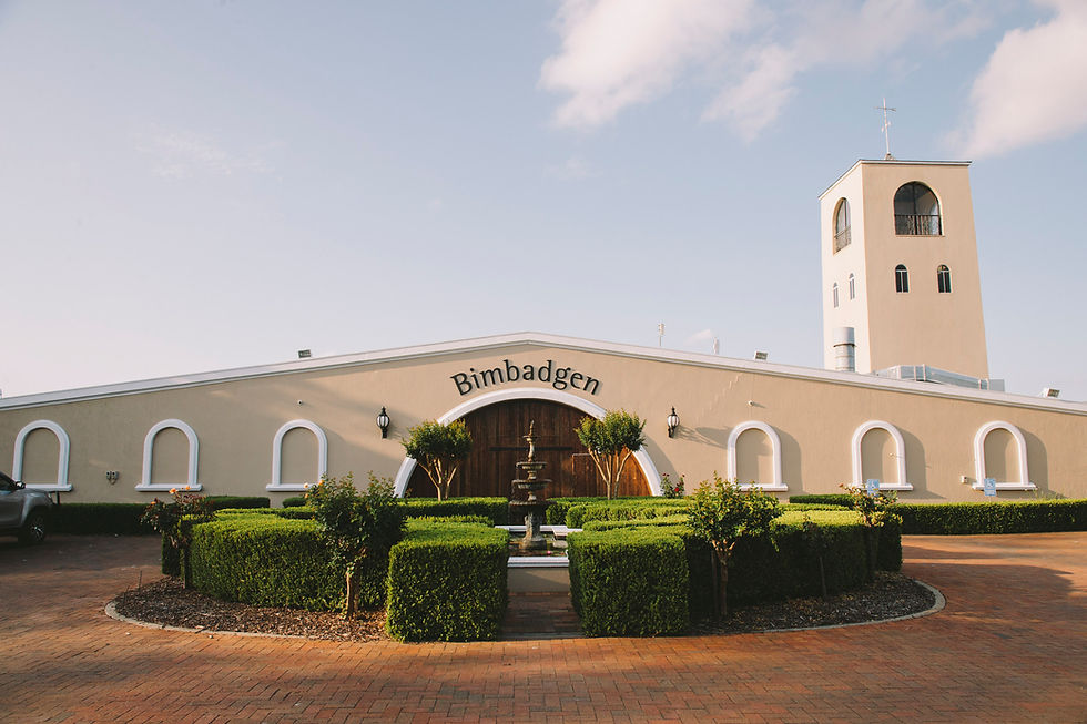 A tan building with "Bimbadgen" text, arched wooden door, and a tower. Green hedges and a fountain in front, under a clear sky.