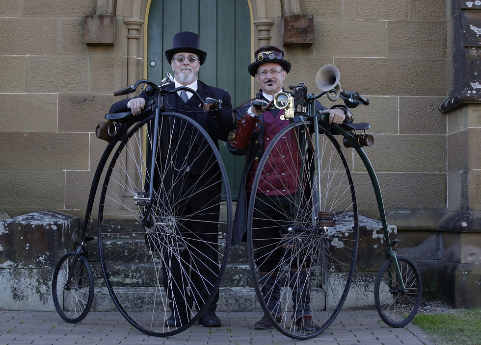 Two men in vintage attire with top hats pose with antique penny-farthings in front of a stone building with a green door, exuding a nostalgic charm.