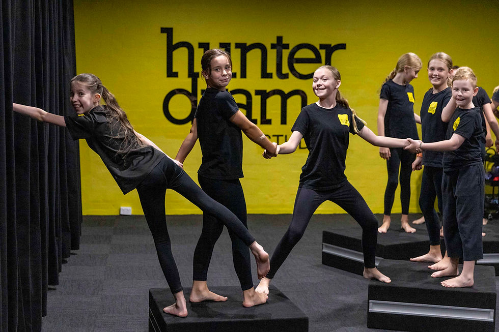 Children in black outfits smile and balance on blocks in a yellow-walled studio labeled "hunter drama". They're holding hands, creating a playful mood.