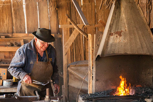 Elderly blacksmith in hat and apron works beside a glowing forge in a rustic wooden workshop.