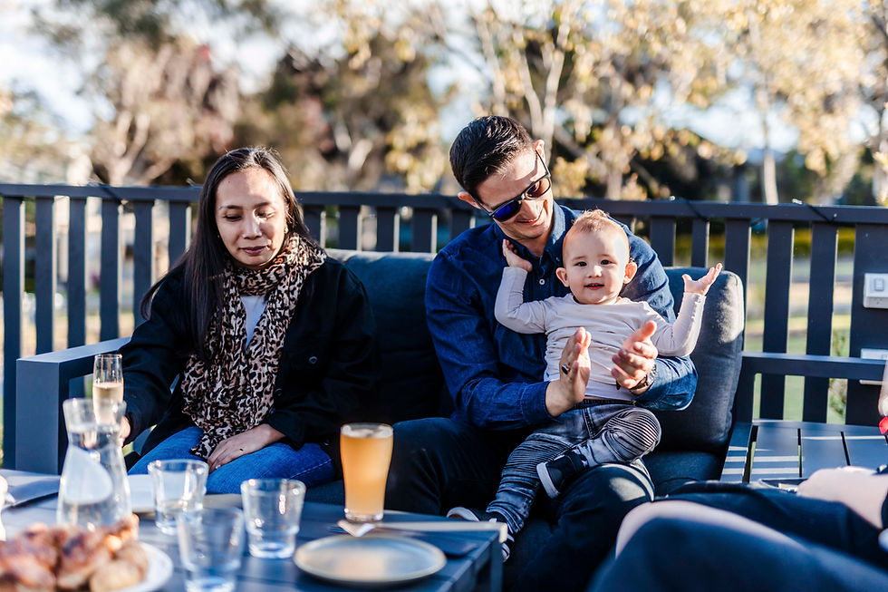 Man holding a smiling baby, seated on a patio with a woman. Drinks and pastries on a table. Sunny outdoor setting with trees.