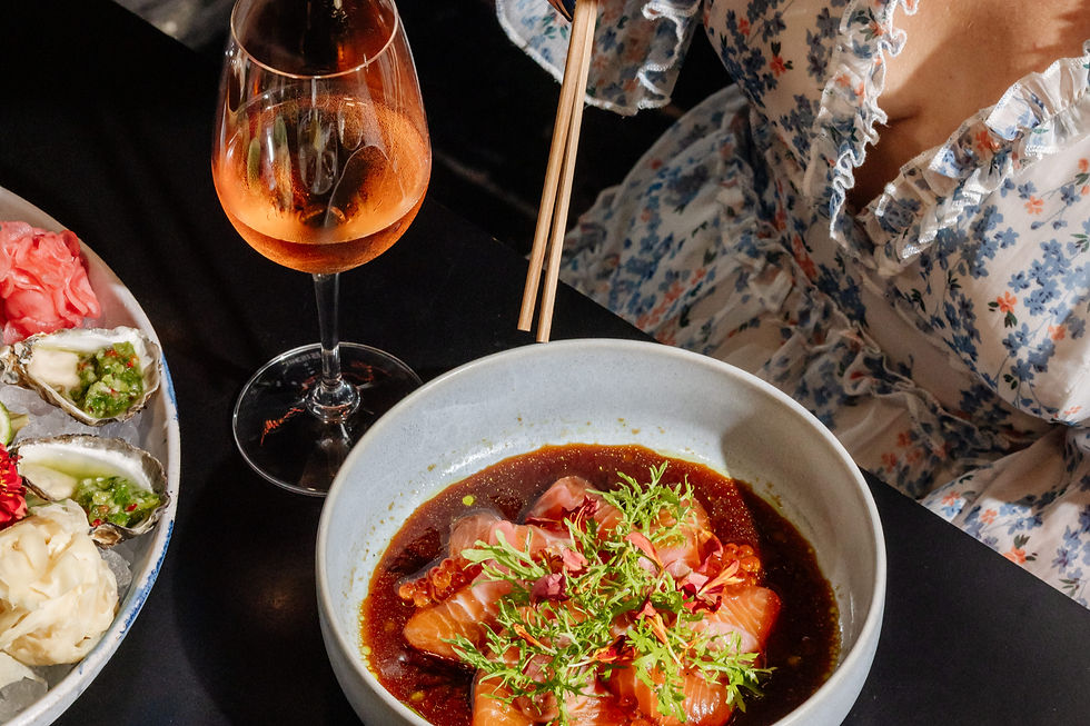 Elegant seafood meal on a dark table: sashimi bowl, oysters, and a rosé drink beside a floral dress, creating a chic, romantic mood