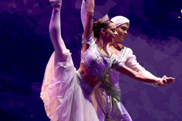 Ballet dancers in ornate costumes perform on stage; woman in arabesque, man supports her. Purple-lit backdrop, elegant and focused mood.